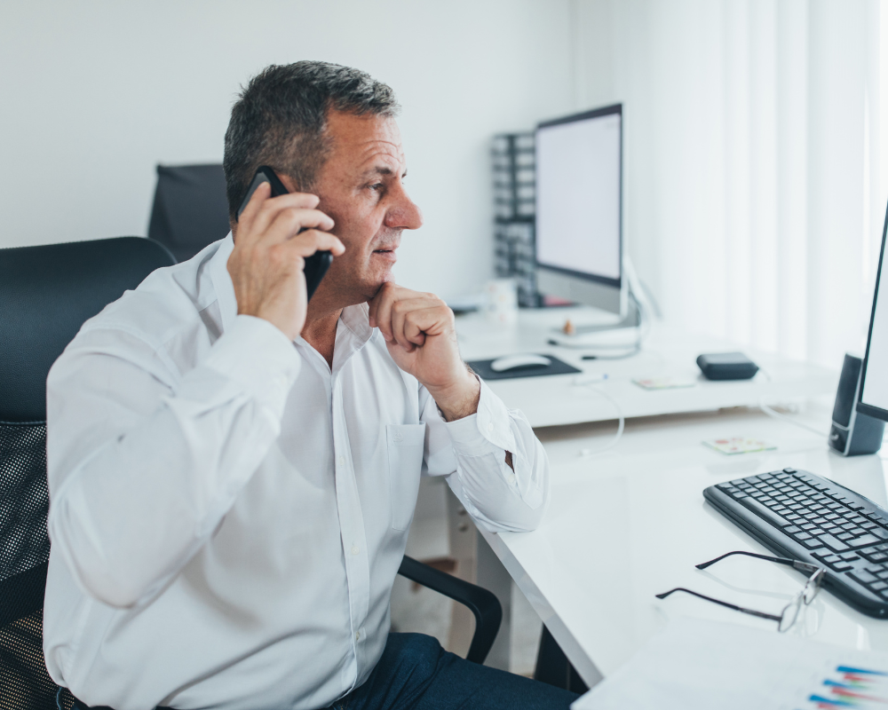Mature businessman in a white shirt talking on the phone at his desk with a computer.