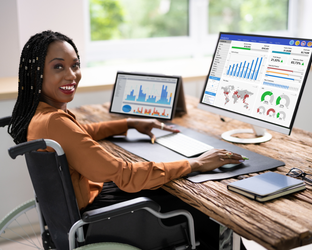 Smiling woman in a wheelchair working at a desk with two monitors displaying data charts and graphs.