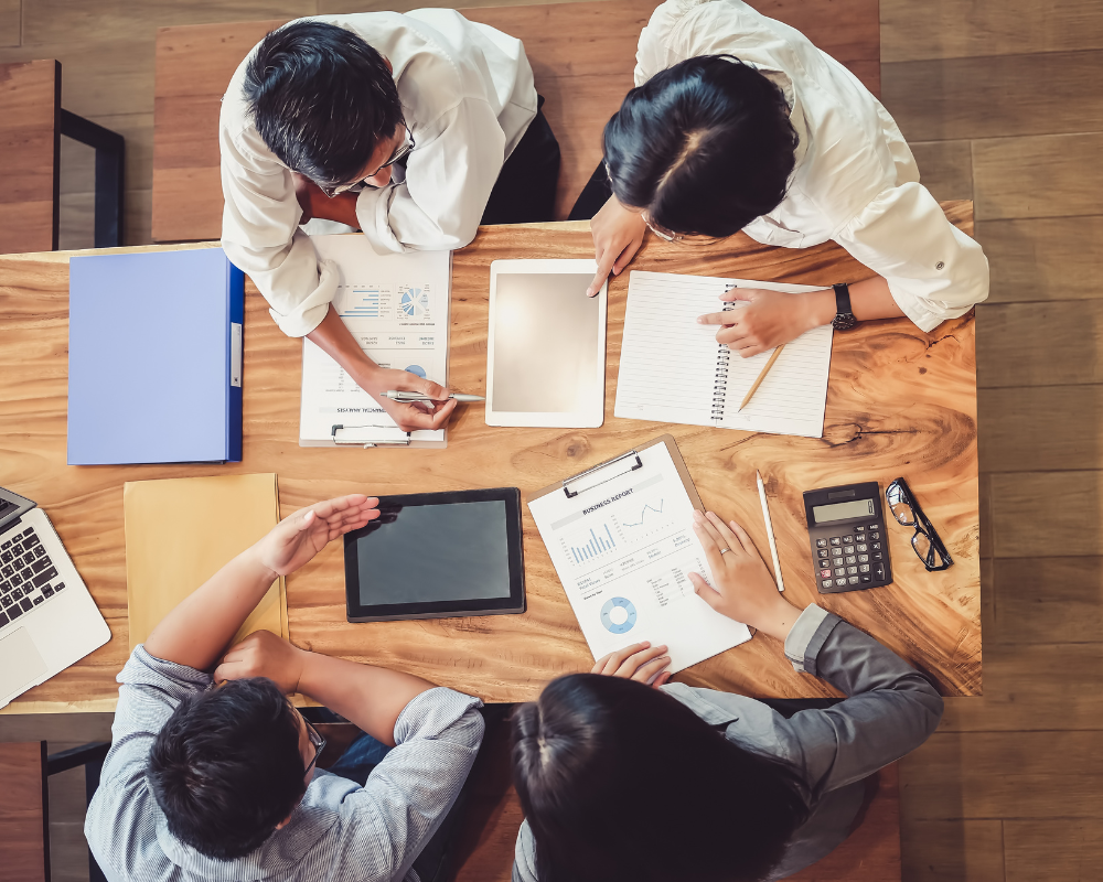 Overhead view of four colleagues working together at a wooden table with charts, notebooks, and tablets.