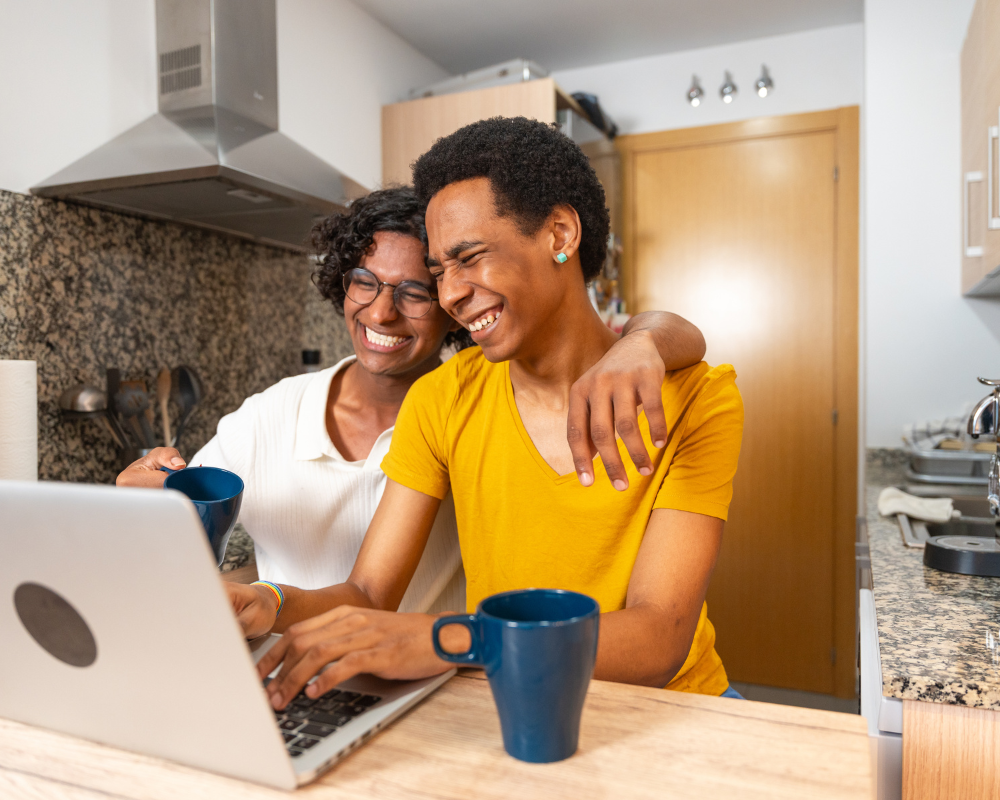 Happy couple sitting in the kitchen with coffee mugs, smiling while looking at a laptop.