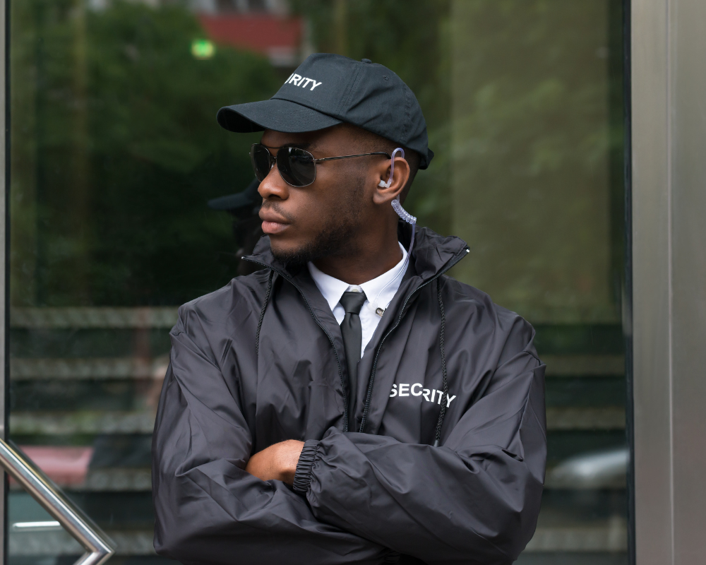 Security guard in black jacket, cap, and sunglasses standing with arms crossed outside a glass building