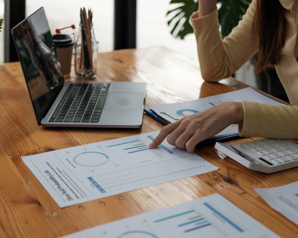 Person pointing at charts and graphs on printed reports next to a laptop.