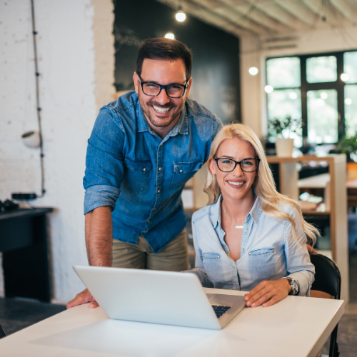 Man and woman wearing glasses smiling while working together at a laptop in a modern office.