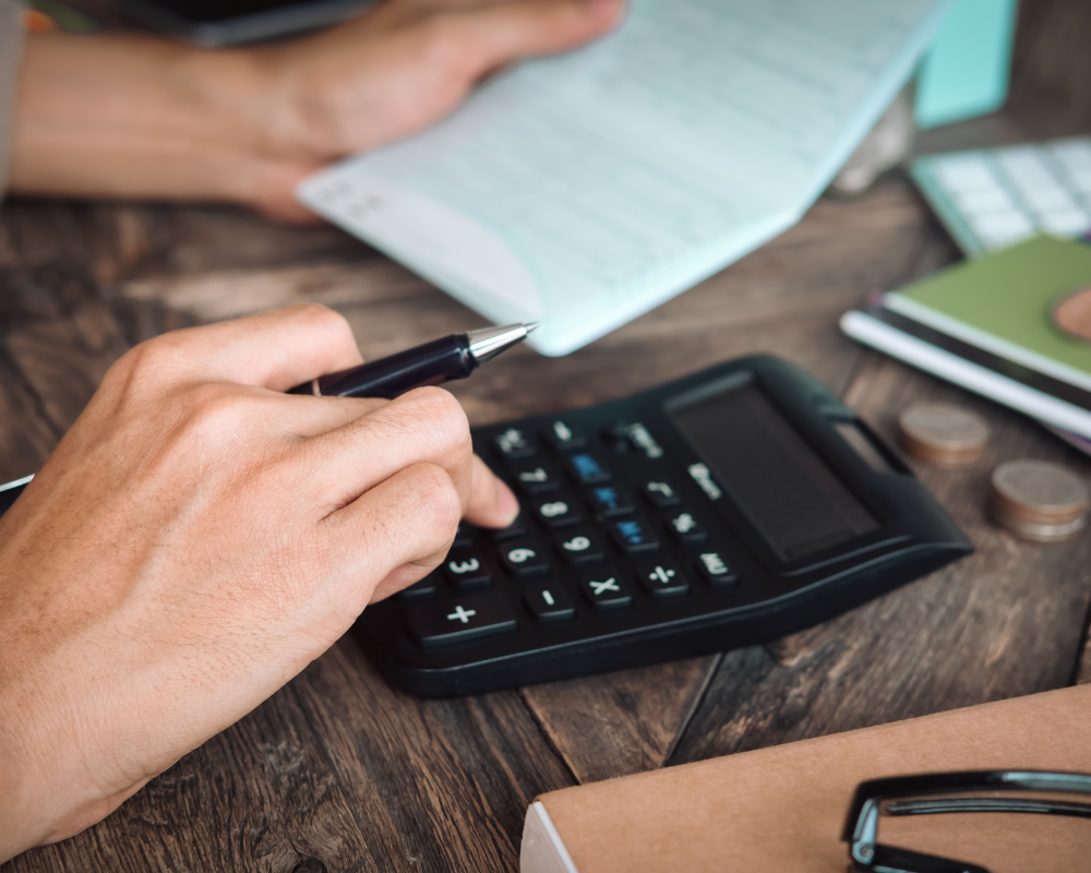 Close-up of hands using a calculator with coins and notebook on a wooden desk.