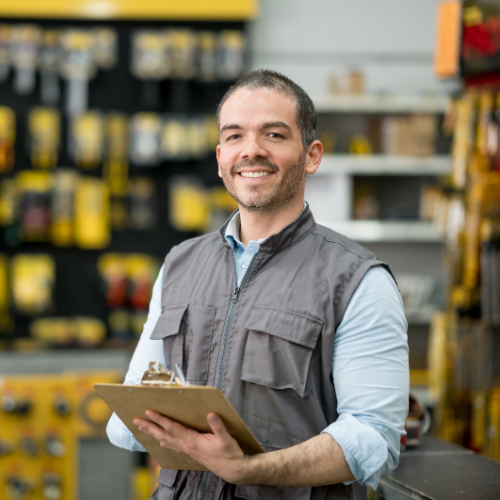 Smiling man in a hardware store holding a clipboard, standing in front of shelves of tools.