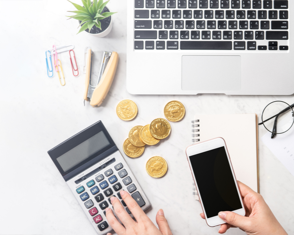 Top view of desk with laptop, smartphone, calculator, gold coins, and office supplies.