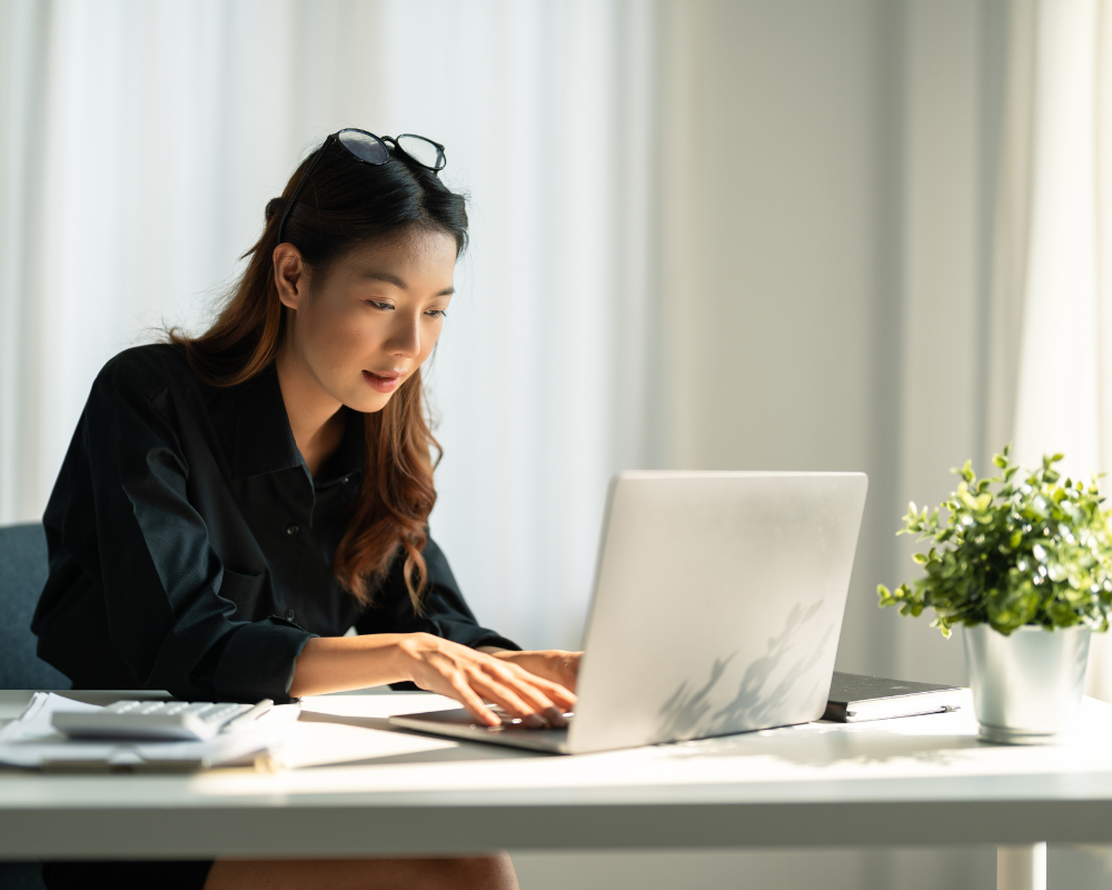 Young woman in a black shirt working on a laptop at a desk with a small plant.
