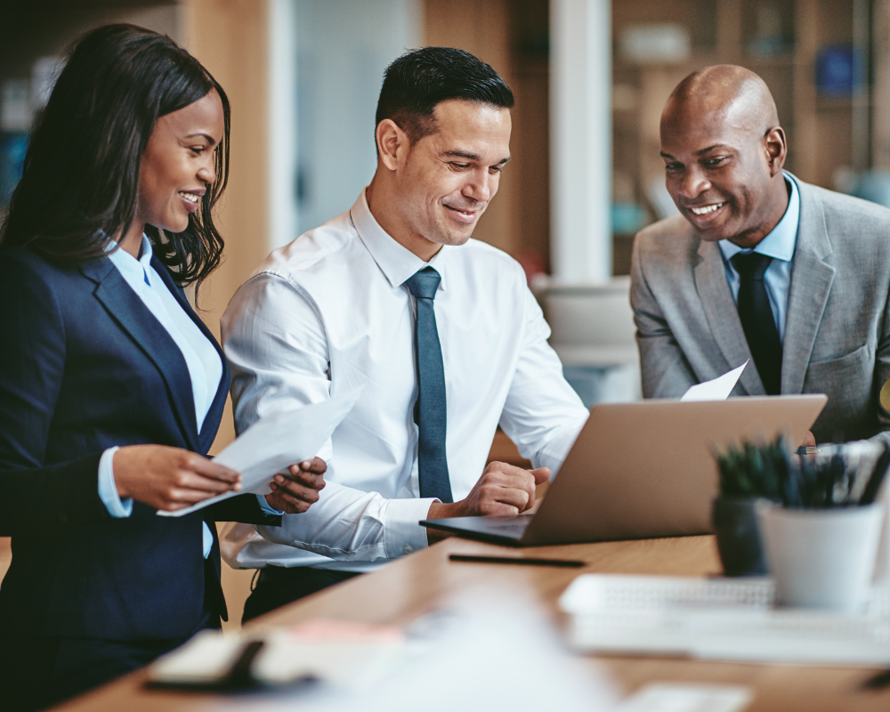 Three business professionals in suits smile and look at documents while gathered around a laptop in a modern office setting.
