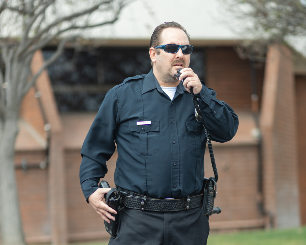 Security officer wearing sunglasses and dark uniform speaking into a handheld radio outside a building