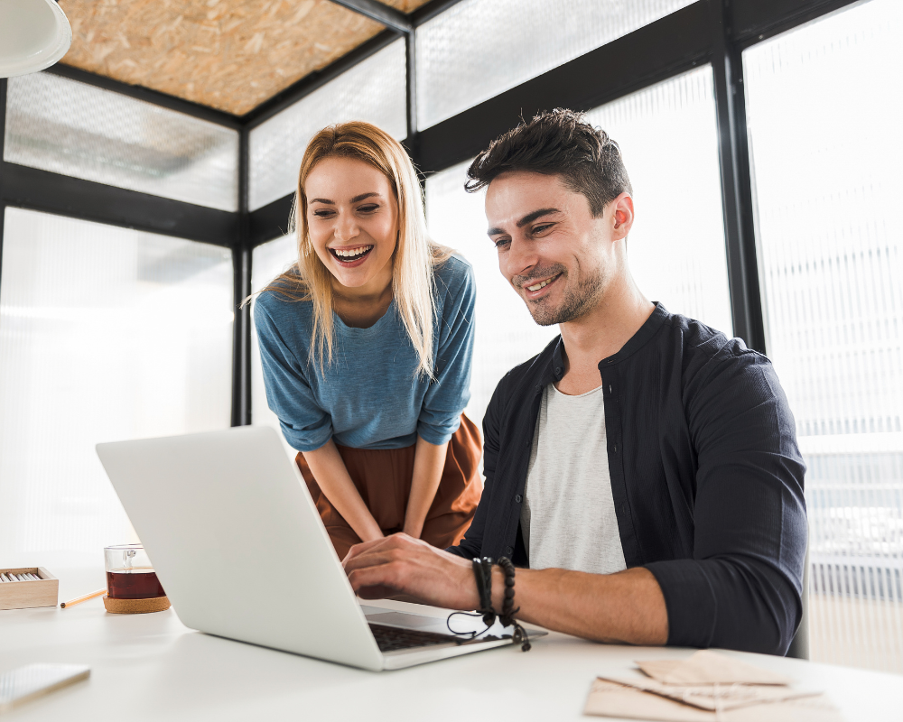 Two colleagues smiling and working together on a laptop at a modern office desk.
