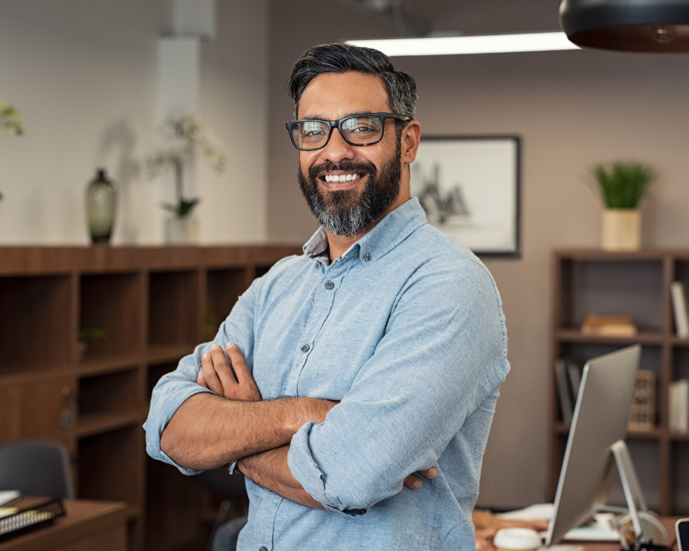 Confident man with glasses standing in an office, smiling with arms crossed.