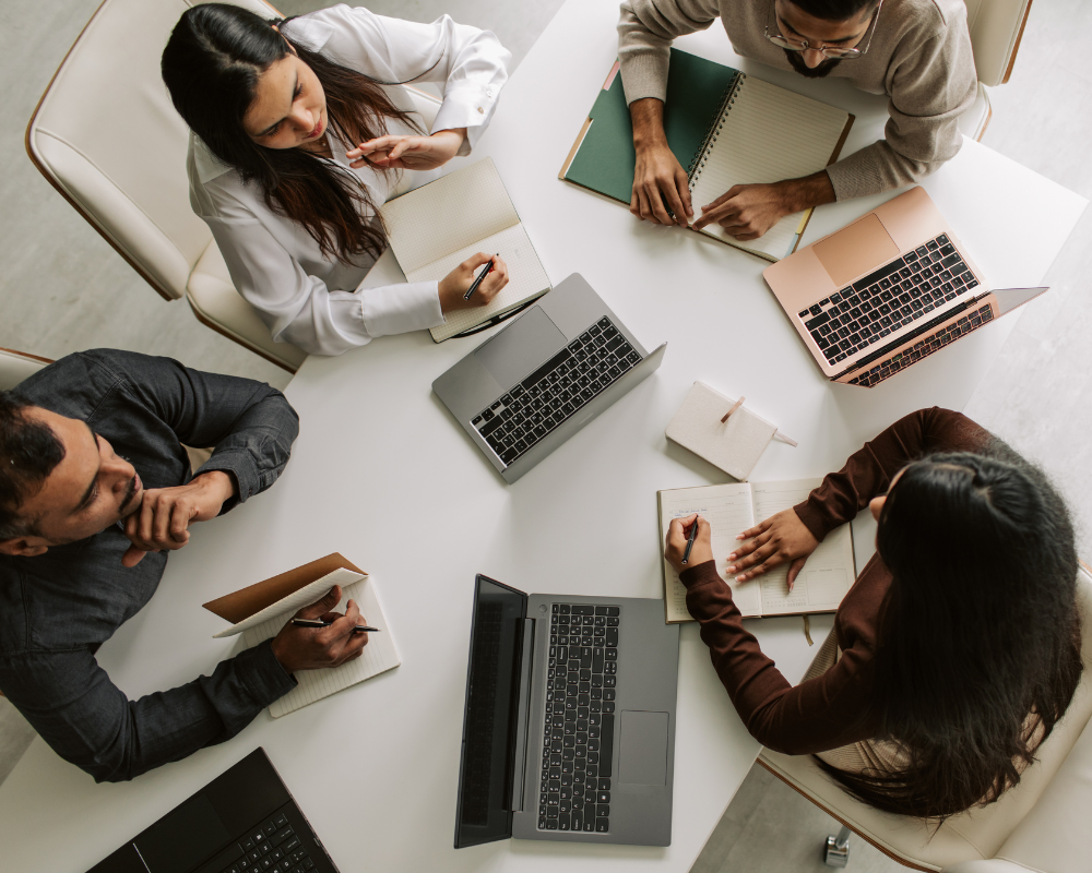 Overhead view of four people seated at a table with laptops and notebooks, collaborating in a meeting.