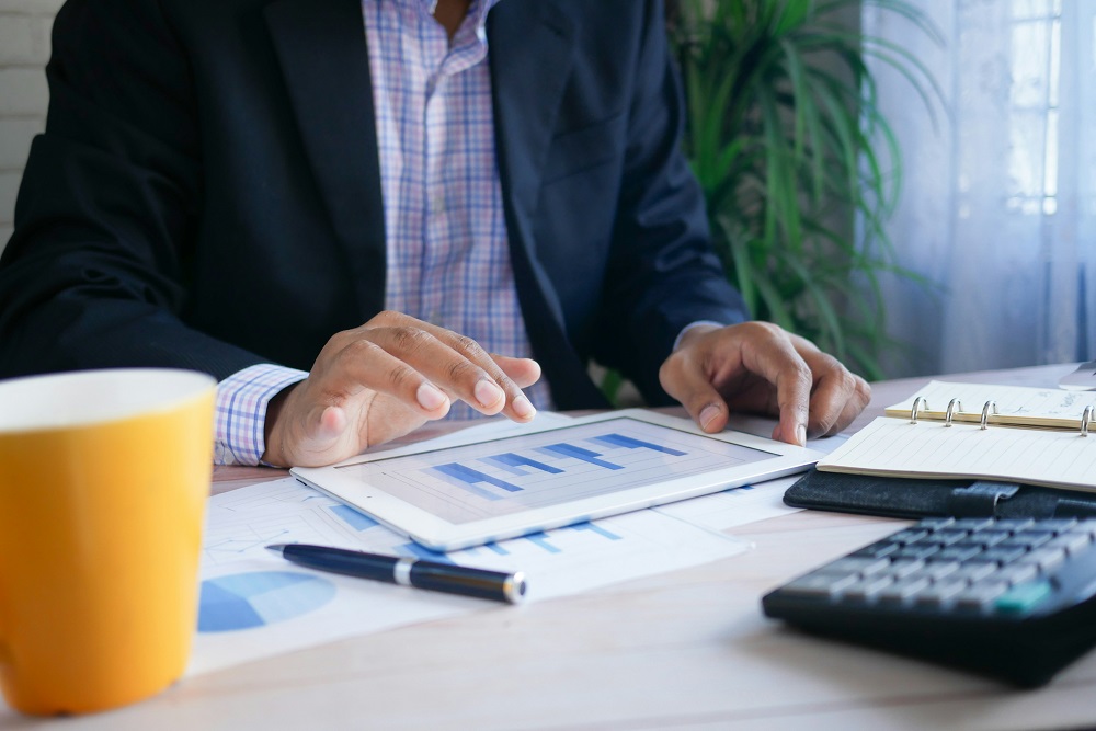 Businessman in a suit reviewing bar chart data on a tablet at a desk with a calculator, notebook, and coffee mug