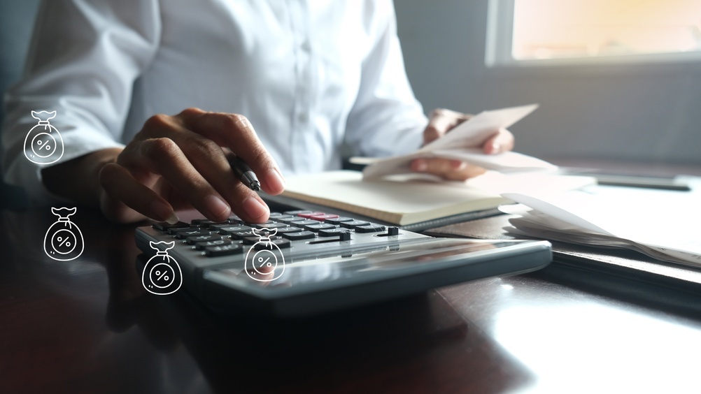 A close-up shot of a person using a calculator at a desk while holding papers, featuring illustrated graphic icons of money bags with percentage symbols hovering in the foreground.