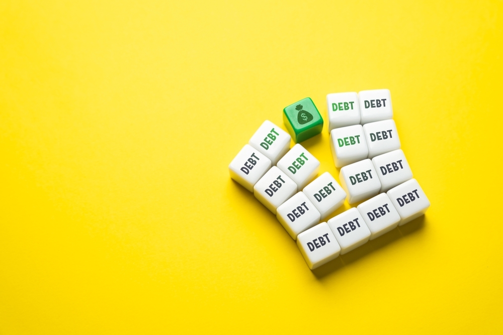 White blocks labeled "DEBT" stacked on a bright yellow background next to a single green block with a currency icon.
