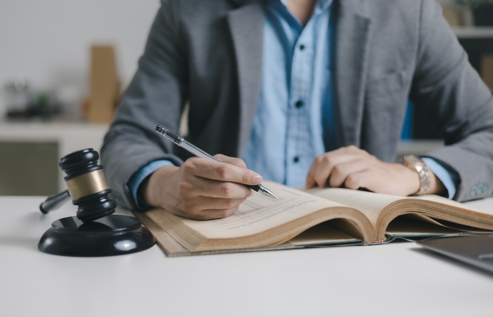 A person in a gray blazer writing with a pen in an open legal book next to a black wooden gavel on a desk.